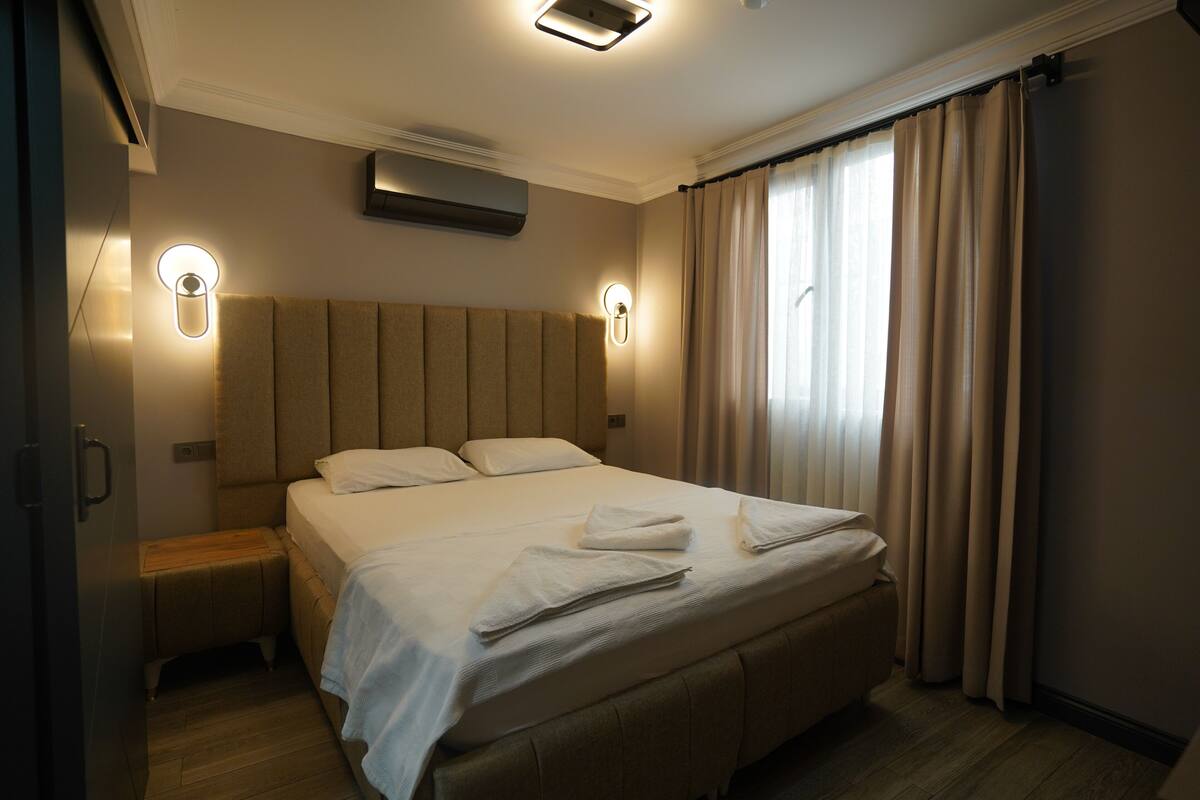 A hotel room with a large bed, two bedside tables, a wall-mounted air conditioner, and a window with curtains.