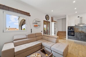 A modern living room with a sectional sofa, a wooden coffee table, and a kitchen area with dark cabinets and a stainless steel range hood.