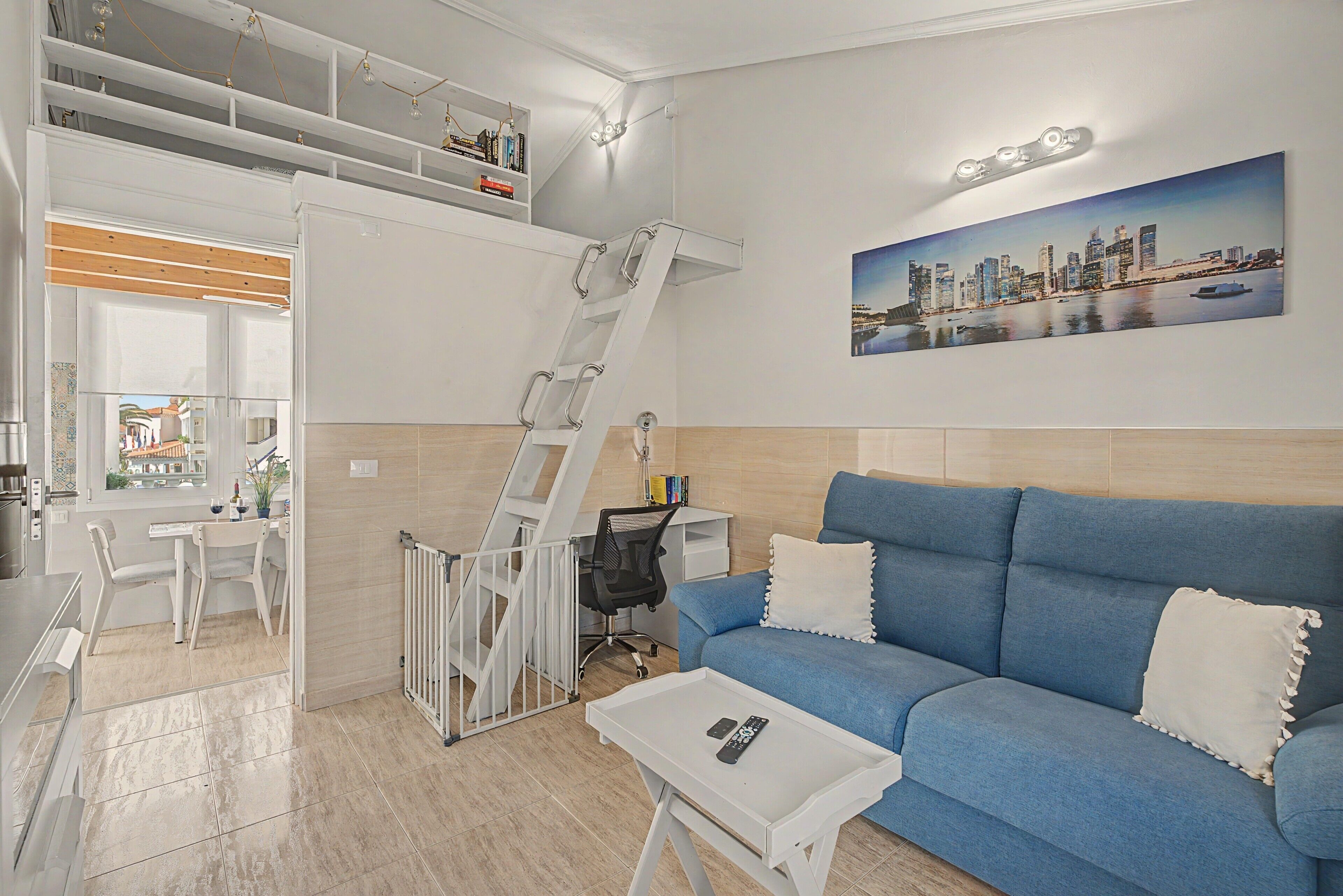 Un salon moderne avec un canapé bleu, une table basse blanche et un tableau représentant un paysage urbain accroché au mur.