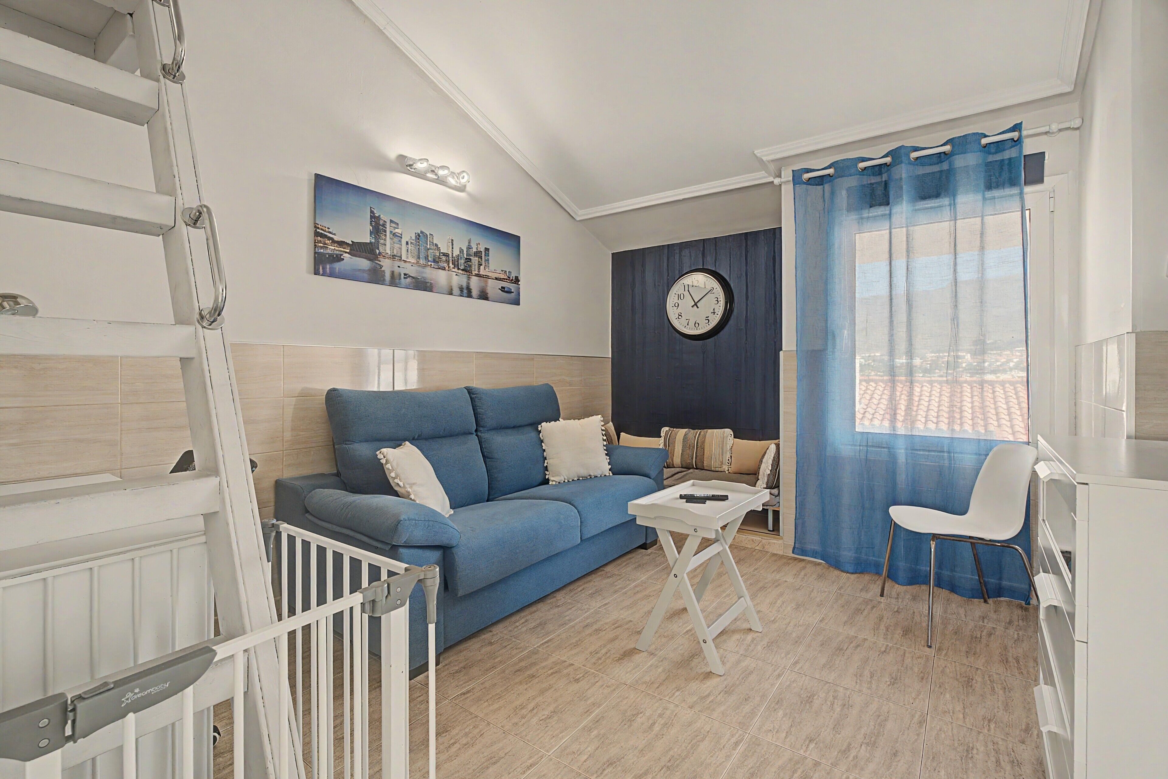Een moderne woonkamer met een blauwe bank, een klein wit tafeltje, een klok en uitzicht op de stad door een raam.