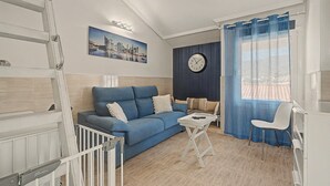 Een moderne woonkamer met een blauwe bank, een klein wit tafeltje, een klok en uitzicht op de stad door een raam.
