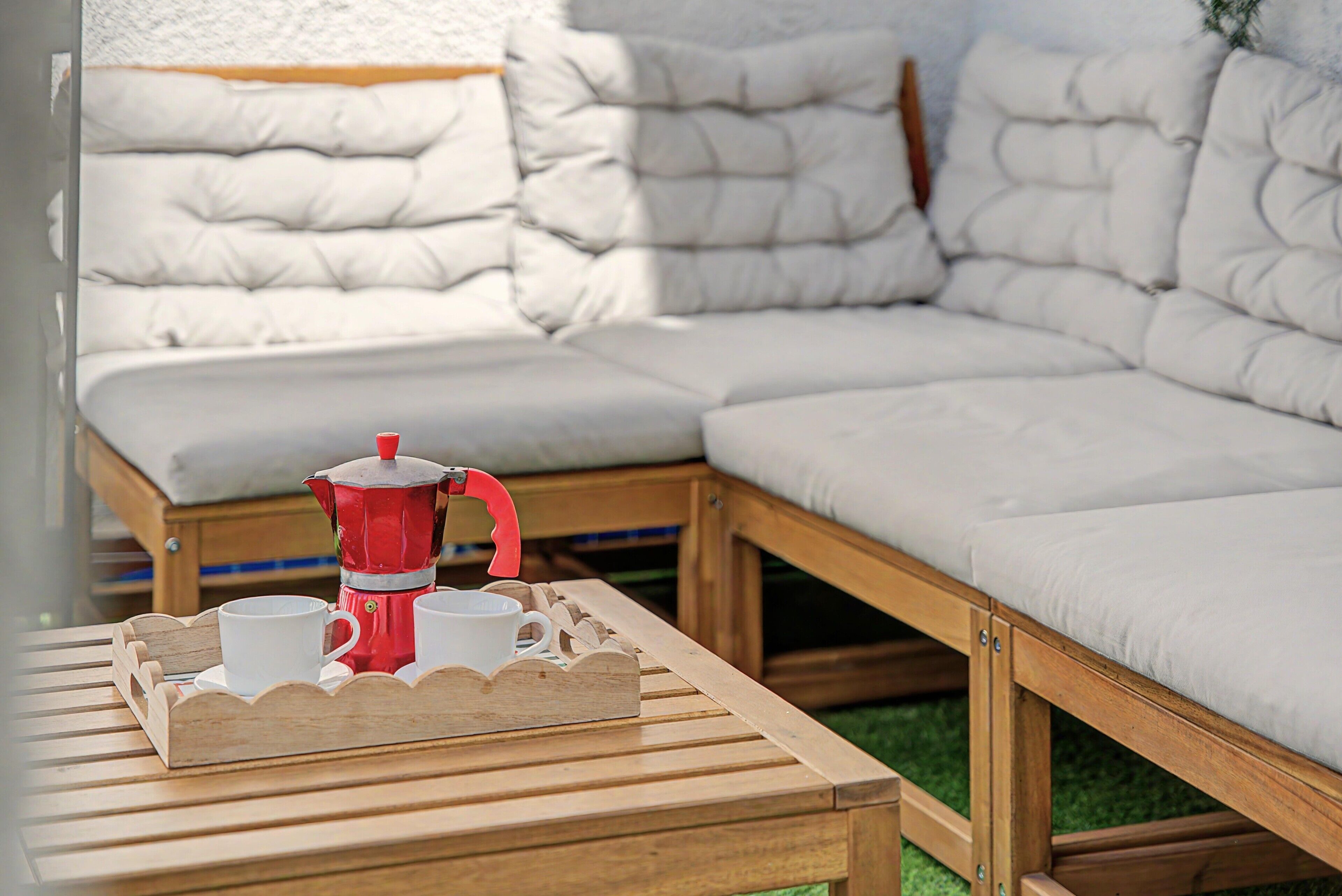 Een houten tafel met een rode koffiepot en witte kopjes, een houten bank en een witte, beklede bank met houten frame.