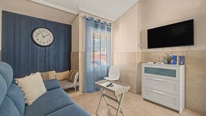 Ein modernes Wohnzimmer mit einem blauen Sofa, einem kleinen weißen Tisch, einem Fernseher und einer Kommode mit Gegenständen darauf.