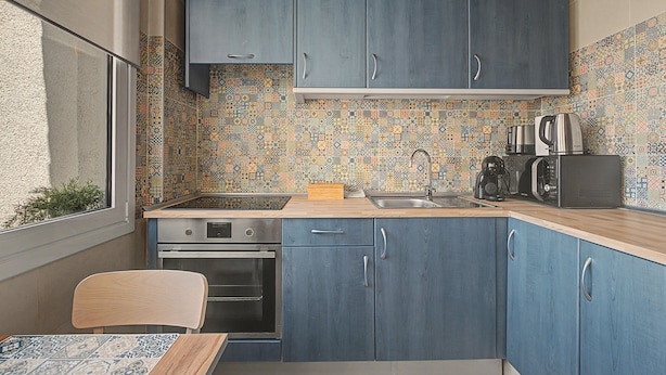 Een compacte keuken met blauwe kastjes, een betegelde spatwand en moderne apparatuur, waaronder een fornuis, oven en magnetron.