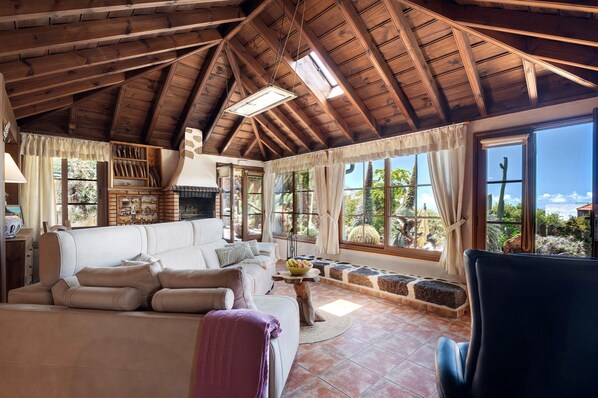 A cozy living room with a high wooden ceiling, a fireplace, a sofa, and large windows offering a view of the outdoors.
