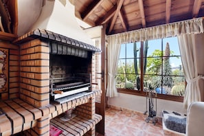 A brick-built outdoor kitchen with a chimney, a window with curtains, and a view of cacti and trees.