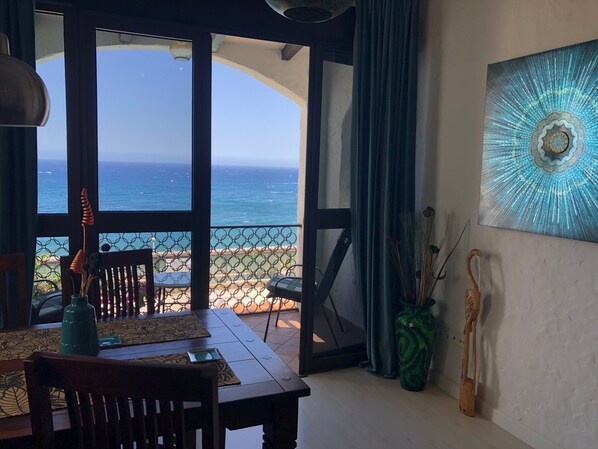 Une pièce avec vue sur l’océan, une table à manger avec des chaises, un vase contenant des plantes séchées et un élément décoratif mural.