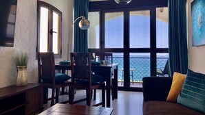 A room with a dining table, chairs, a sofa, a hanging light fixture, and a view of the sea through large windows. - Holiday Home 'Panoramic Ocean View' with Shared Pool, Wi-Fi and Air Conditioning (Playa del Inglés)