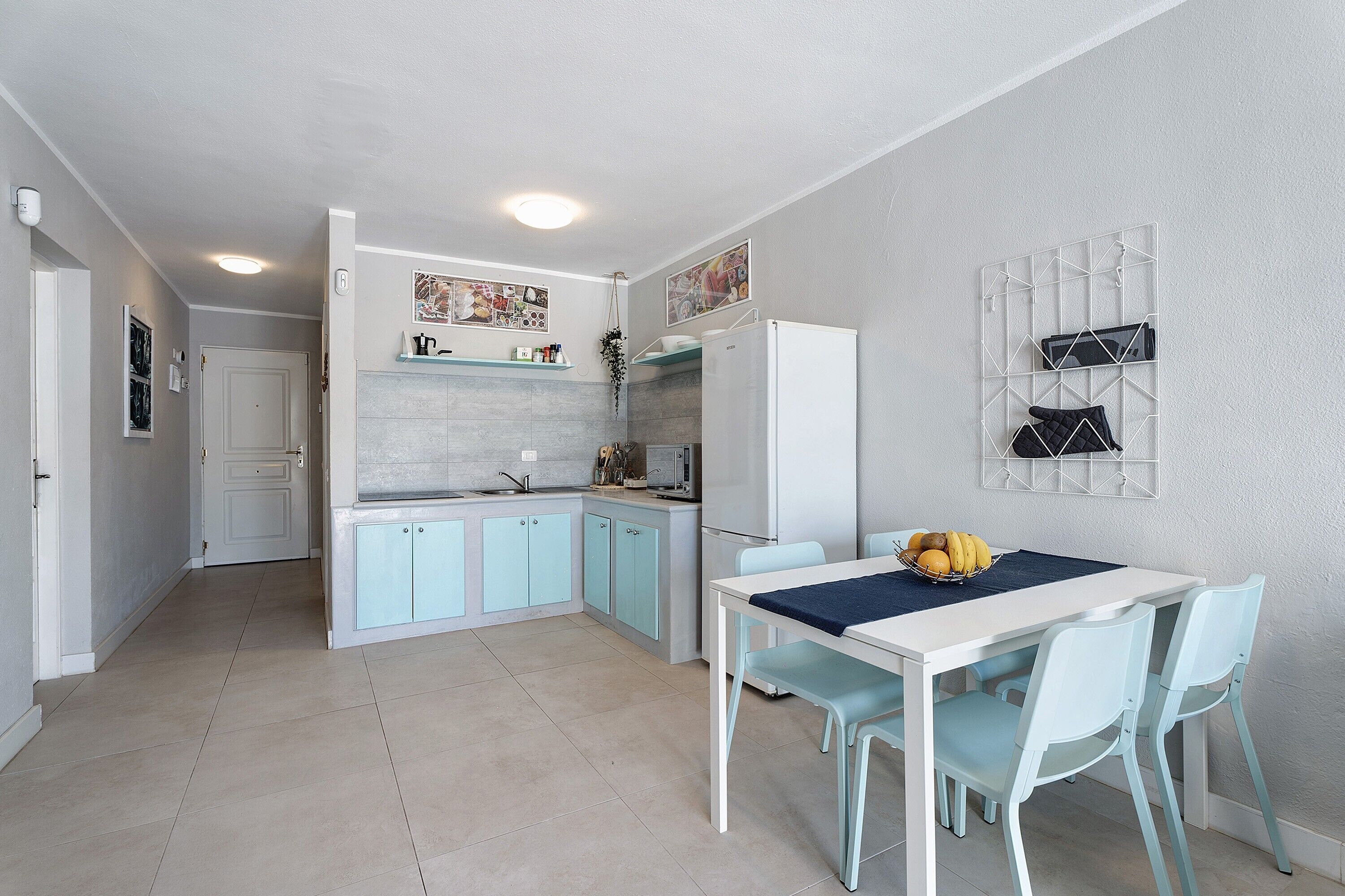 Een kleine keuken met lichtblauwe kastjes, een witte koelkast en een eethoek met een tafel en stoelen.