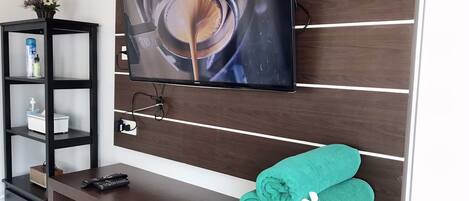 A modern living room with a flat-screen TV mounted on a wooden wall, a black entertainment unit with shelves, and a neatly rolled green towel on a wooden table.