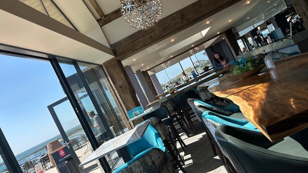 A modern restaurant with large windows overlooking the beach, wooden tables and blue cushioned chairs, and a unique chandelier. - Clydfan No 1 Trearddur Road (Trearddur Bay)