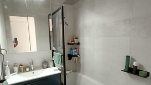 Un baño con lavamanos blanco, espejo y bañera. Hay repisas con artículos de tocador y una alfombra verde en el suelo.