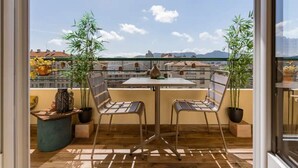 Un balcón con mesa y sillas, plantas en macetas y vistas a edificios y montañas.