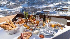 Un buffet de petit-déjeuner proposant une variété de viennoiseries, de fruits et de boissons, le tout disposé sur une table avec une vue sur les montagnes en arrière-plan.