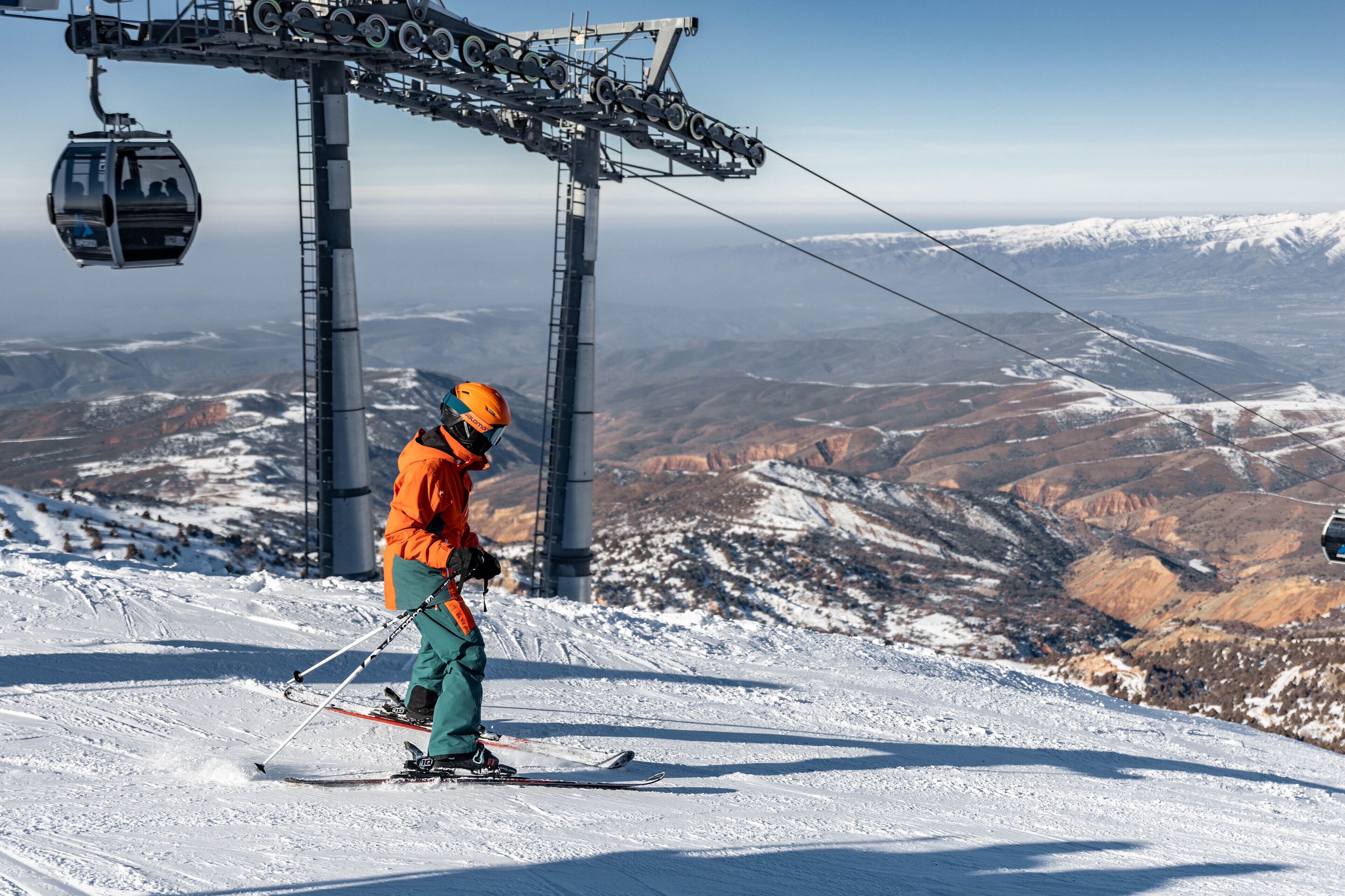 Eine Seilbahn mit Gondeln, eine Person in orangefarbener und grüner Skileidung und eine verschneite Berglandschaft.