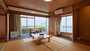 Una habitación con esteras de tatami, una mesa con toallas, sillas, un calentador y ventanas grandes con cortinas.