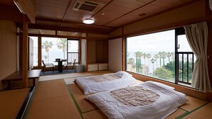 Una habitación con tatamis, cama con ropa de cama blanca, balcón con mesa y sillas, y vistas a palmeras y al mar.