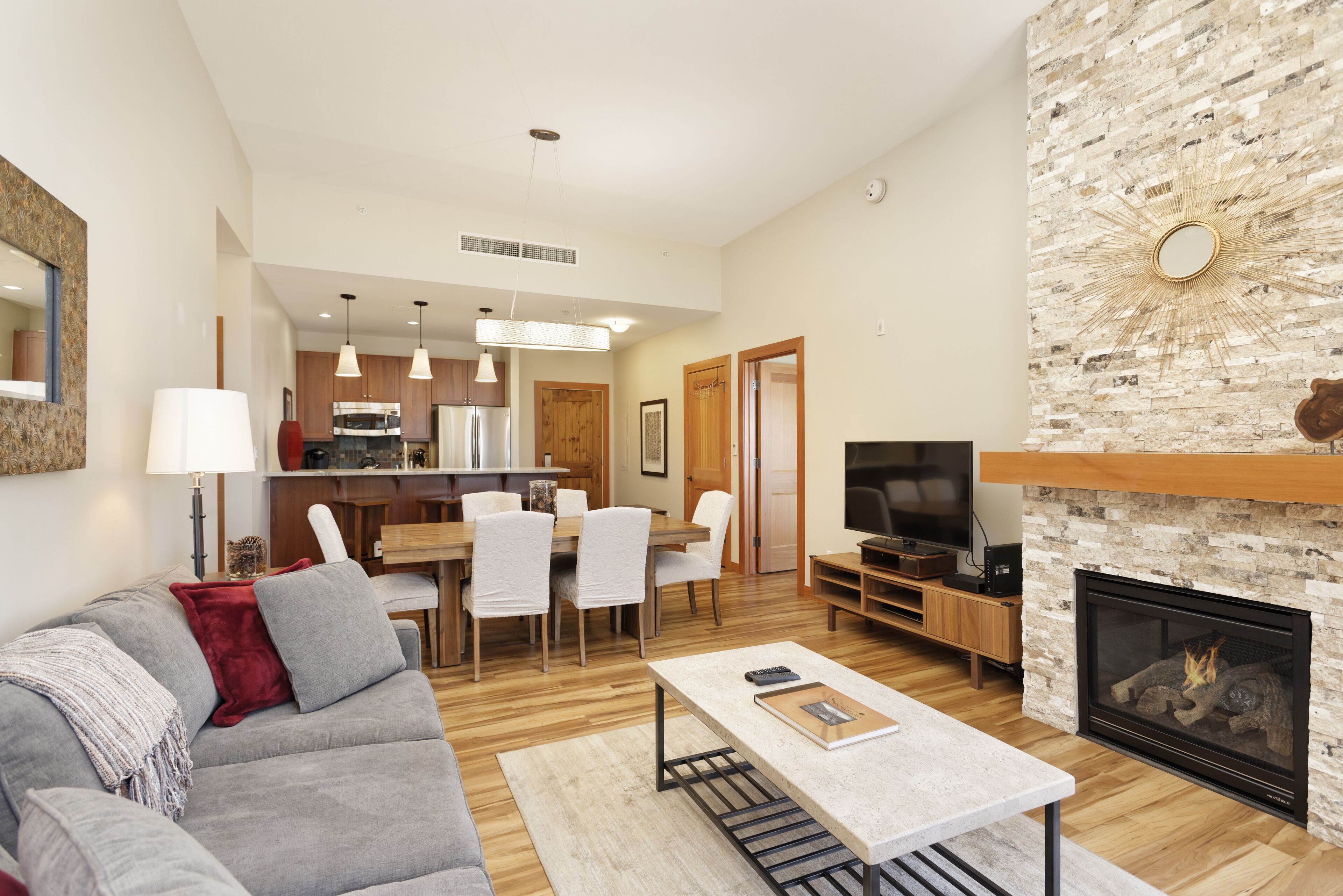 A modern living room with a gray sofa, a coffee table, and a fireplace. There is a dining area with a wooden table and chairs, and a kitchen in the background.