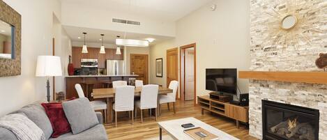 A modern living room with a gray sofa, a coffee table, and a fireplace. There is a dining area with a wooden table and chairs, and a kitchen in the background.