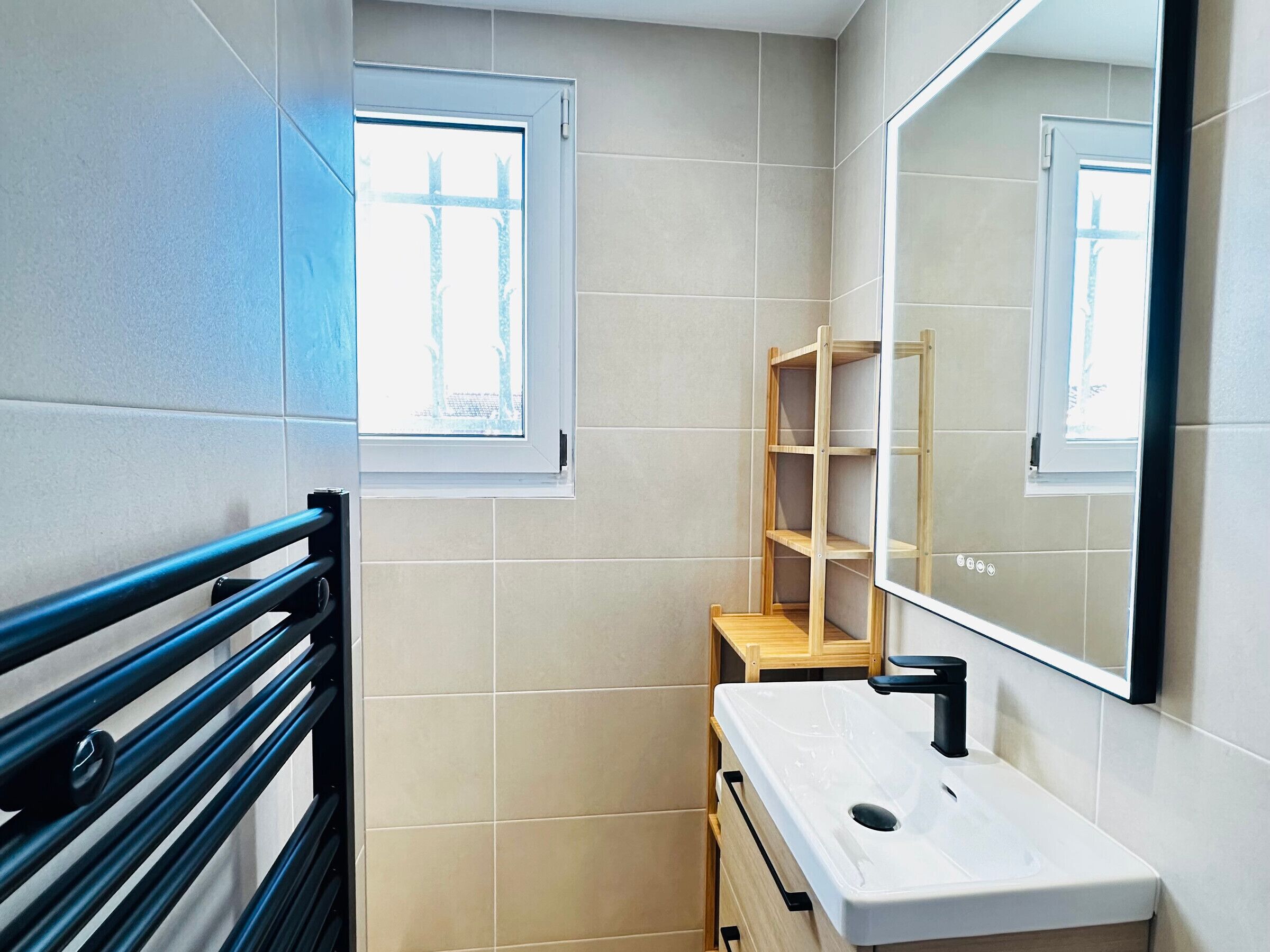 Un bany amb un lavabo blanc, un mirall, una finestra i una prateleira de fusta.