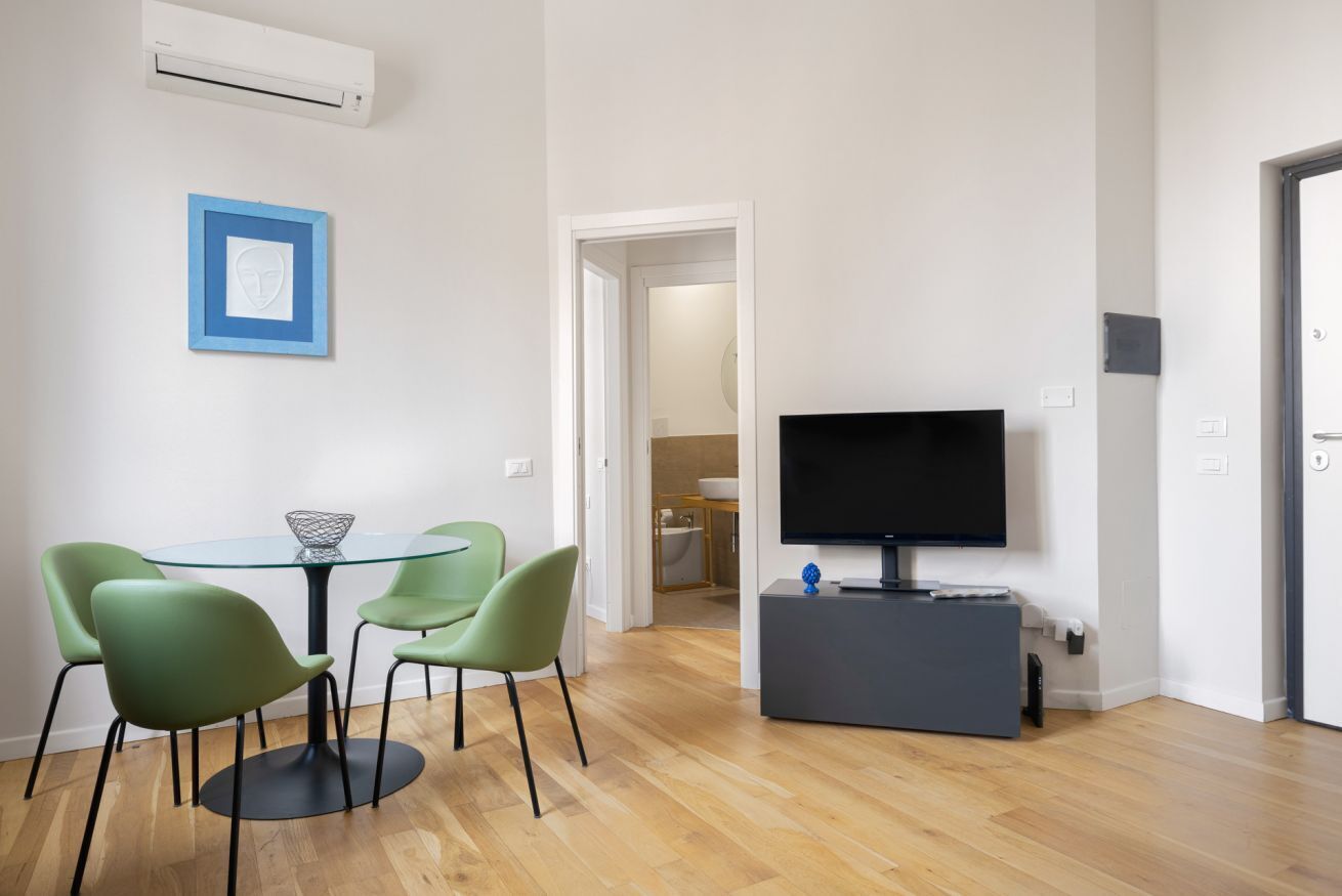 Una sala de estar moderna con una mesa redonda de vidrio, sillas verdes, un televisor de pantalla plana sobre un soporte gris y un cuadro en la pared.