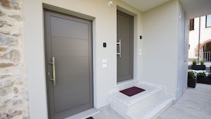 A modern building entrance with a stone wall, a grey door, and a small set of steps leading to a door with a red mat.