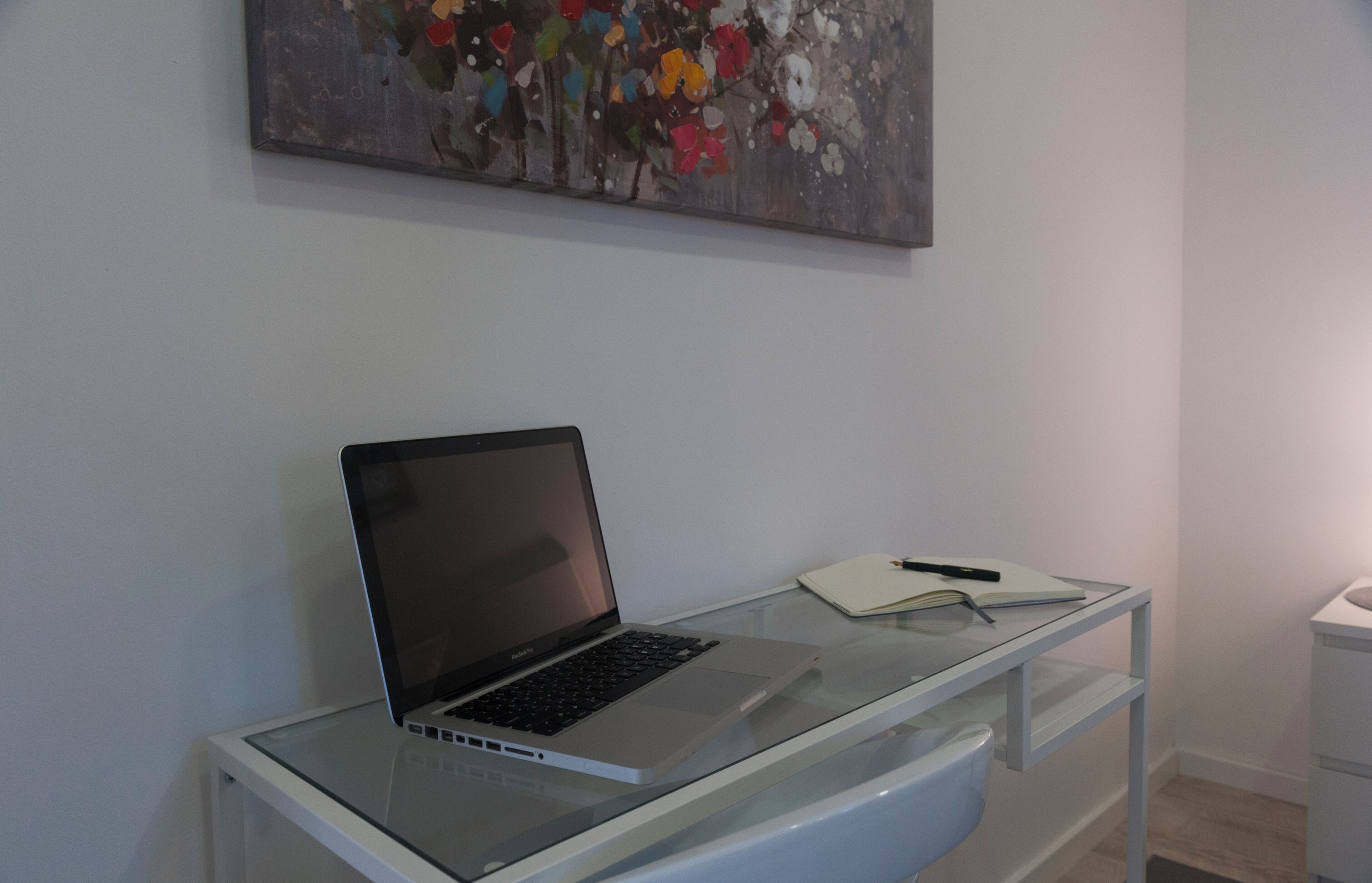 Ein Laptop steht auf einem Glastisch, daneben liegen ein Notizbuch und ein Stift. An der Wand hängt ein Gemälde, im Hintergrund ist ein weißes Schranksicht.