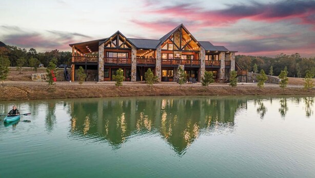 A large, multi-story lodge with a stone facade and large windows, situated by a calm lake.