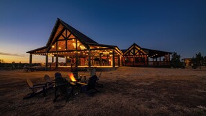 A lodge with a large open patio area featuring Adirondack chairs and a fire pit, surrounded by wooden structures with string lights.