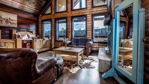 A rustic log cabin interior with a large wooden dining table, leather sofa, and a cozy fireplace.