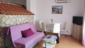 Ein Wohnzimmer mit einer Steinsäule, einem violetten Sofa, einem Glastisch mit Stühlen, einem Fernseher und einer Klimaanlage.