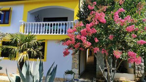 Ein zweistöckiges Gebäude mit gelber Fassade, einem Balkon mit weißen Brüstungen und einem Garten mit rosa Blumen und Agavenpflanzen.