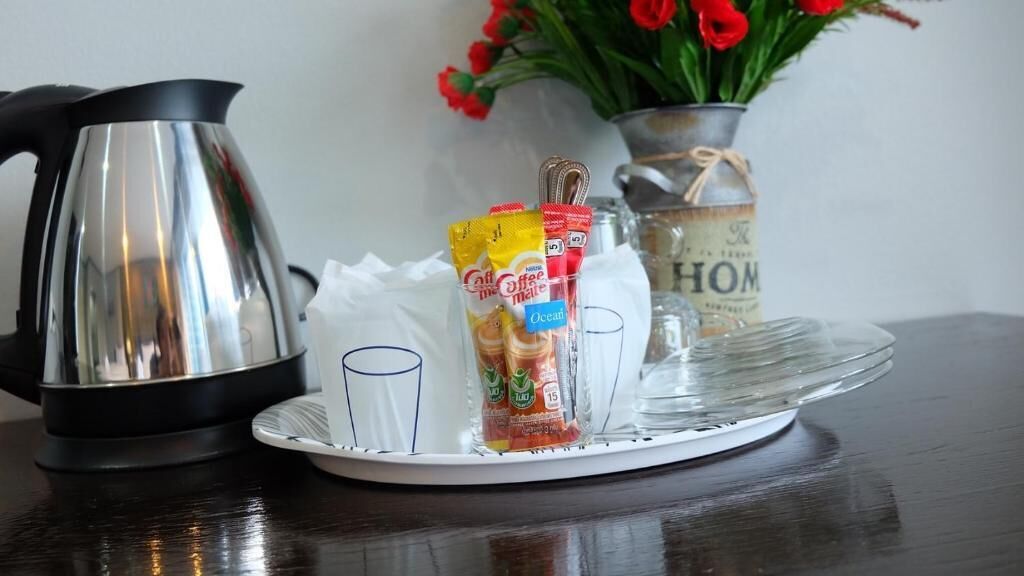 Una tetera de acero inoxidable, una bandeja de vidrio con sobrecitos de café y un florero con flores rojas sobre una mesa de madera.