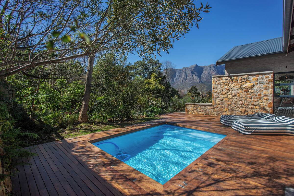 Piscina com deck de madeira, rodeada por vegetação e um muro de pedra, com montanhas ao fundo.