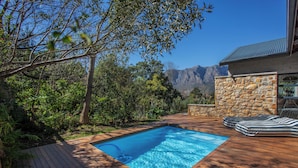 Una piscina con muelle de madera, rodeada de vegetación y un muro de piedra, con montañas al fondo.