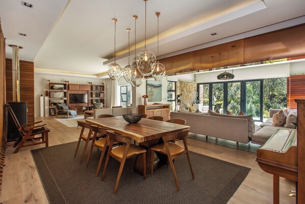 A spacious kitchen with a large wooden table and chairs, a hanging light fixture, and a view of the outdoors through large windows.