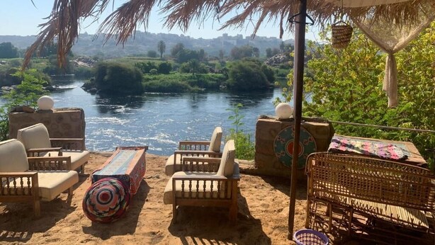 A riverside seating area with wicker furniture and a colorful rug, overlooking a river and greenery. - Nubian Farm  (Aswan)