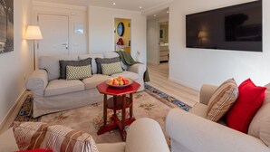 Un salon avec un canapé, des fauteuils, une table basse sur laquelle se trouvent des fruits, une lampe et un téléviseur fixé au mur.