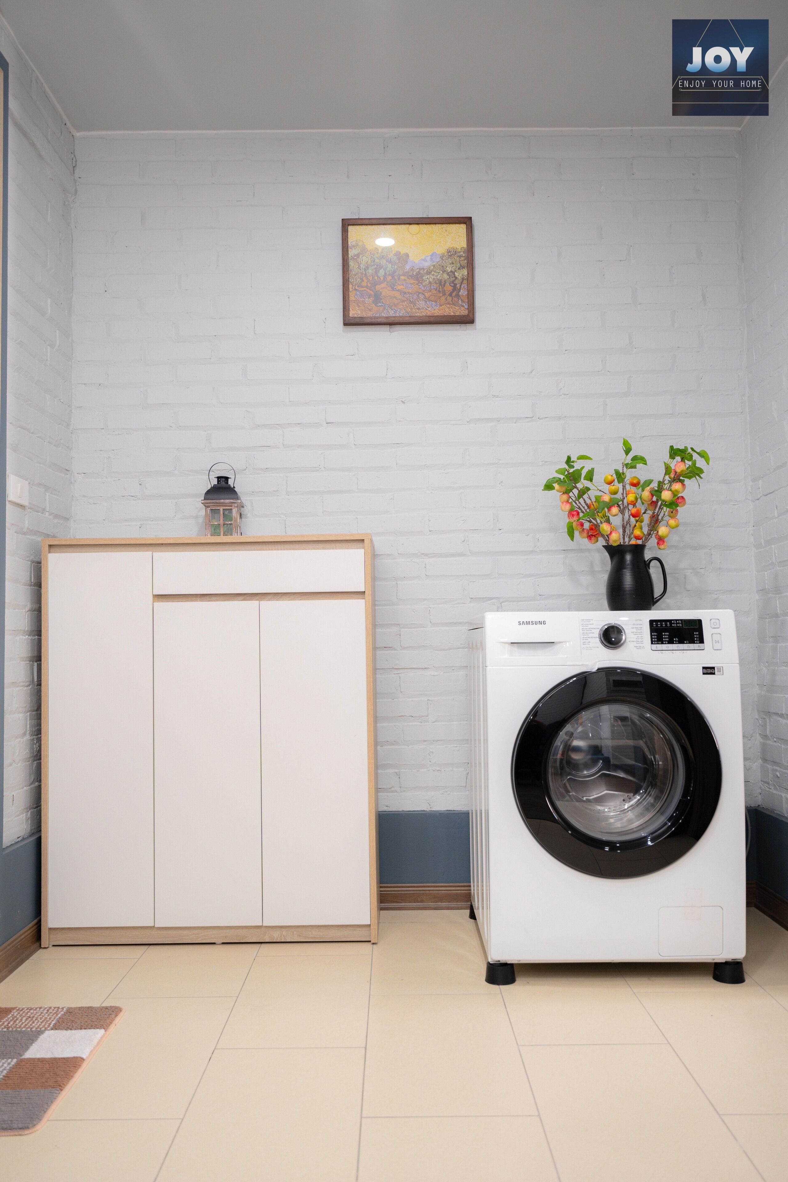 A laundry room with a white brick wall, a Samsung washing machine, a cabinet, and a small table with a vase of flowers.