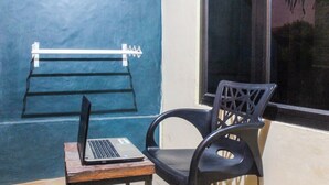 A laptop on a small wooden table next to a black chair, with a metal rack against a blue wall and a window in the background. - Hotel O We Bro Kost (Nginamanu)