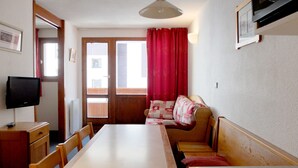 A room with a wooden dining table, chairs, a sofa with a quilt, a television on a stand, a door leading outside, and a window with curtains. - Comfortable Apartment in Tignes, Pet Friendly (Tignes)