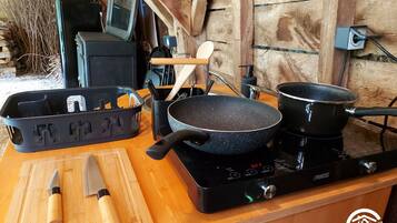Uma cozinha com bancada de madeira, fogĂŁo preto, duas frigideiras, tĂĄbua de corte, faca, cesta de plĂĄstico e colher de pau.