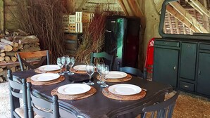 A rustic dining area with a wooden table set for six, surrounded by chairs. A black refrigerator is visible in the background, and a stack of firewood is to the left. - Charming 2-Person Cottage with Garden & Shared Amenities in Montenay (Montenay)