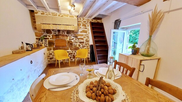 A dining area with a wooden table set for two, a stone wall, and a staircase leading to a loft. - Charming 2-Person Cottage with Garden & Shared Amenities in Montenay (Montenay)
