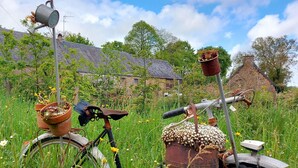 A bicycle with a rusty pot and a watering can attached to it, parked in a grassy field with a house in the background. - Charming 2-Person Cottage with Garden & Shared Amenities in Montenay (Montenay)