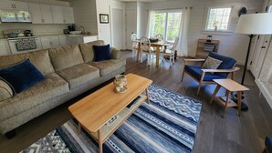 A cozy living room with a sofa, coffee table, and patterned rug. A dining area with a table and chairs is visible. The kitchen is in the background with white cabinets and a stove. - Newly renovated waterfront cottage with spectacular sunset views (Sullivan)