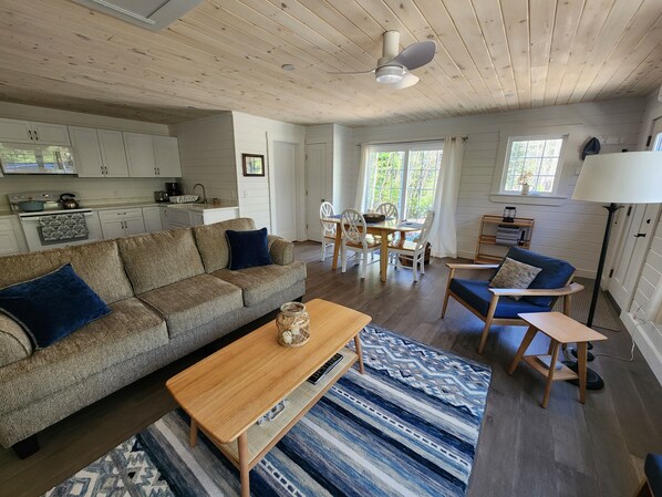 A cozy living room with a sofa, coffee table, and patterned rug. A dining area with a table and chairs is visible. The kitchen is in the background with white cabinets and a stove.