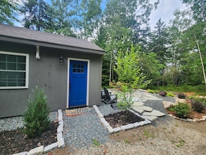 Una casa con puerta azul, un pequeño jardín con un árbol y un patio con muebles de exterior.