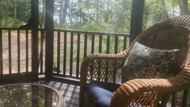 A wicker chair with a cushion on a wooden deck, surrounded by a railing, with a view of a forested area through the railing. - Newly renovated waterfront cottage with spectacular sunset views (Sullivan)