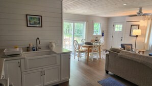 A modern kitchen with white cabinets, a dining area with a wooden table and chairs, and a living room with a sofa and a lamp. - Newly renovated waterfront cottage with spectacular sunset views (Sullivan)